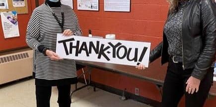 Shows 2 women standing indoors with face masks holding a sign that says Thank You