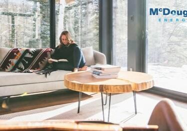 Shows a woman reading a book sitting on a large couch in a large room surrounded by windows