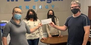 Shows a group of 4 people standing and holding a cheque wearing face masks against wall with McDougall Energy logo