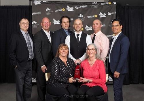 Shows 6 men standing and 2 women sitting in front of them smiling