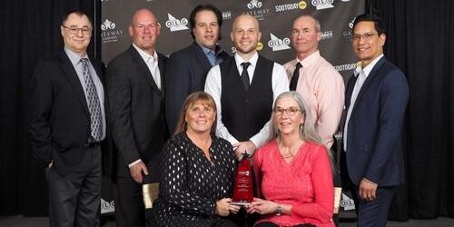 Shows 6 men standing and 2 women sitting in front of them smiling
