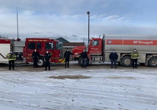 Shows a shot of 7 people outdoors winter with a firetruck and a McDougall fuel truck behind them