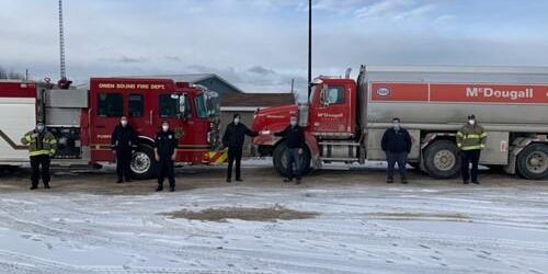 Shows a shot of 7 people outdoors winter with a firetruck and a McDougall fuel truck behind them